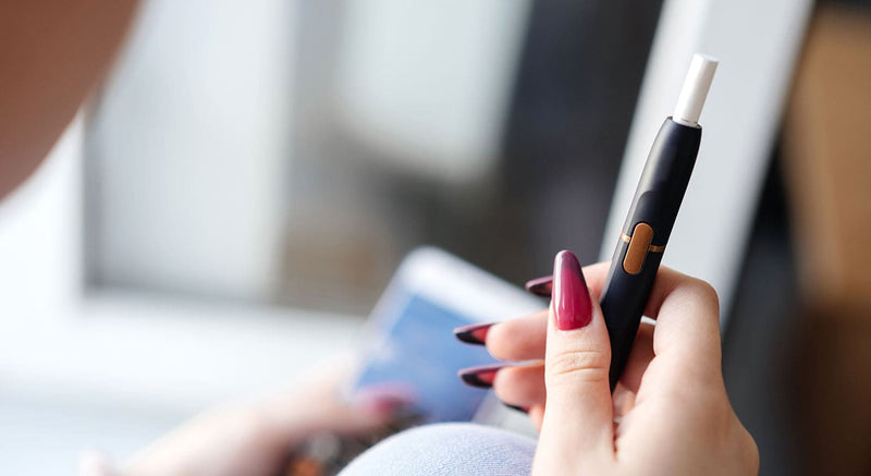 Woman holding heated tobacco device in right hand while scrolling on mobile phone in left hand.